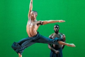 A dancer completing a stag leap in front of another standing dancer and green background.